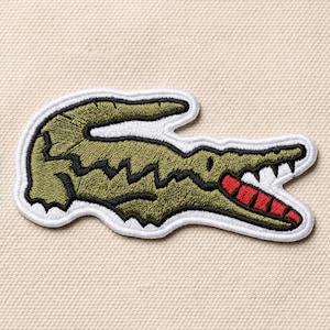 May include: Embroidered patch featuring a green crocodile with a white border. The crocodile has a red mouth and white teeth. The patch is on a beige fabric background. This patch is a classic emblem.