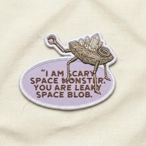 May include: Embroidered patch featuring a cartoon space monster with crystal accents, standing on a purple oval with the text "I AM SCARY SPACE MONSTER. YOU ARE LEAKY SPACE BLOB." The patch is on a cream-colored fabric background.