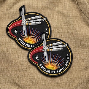 May include: Two embroidered patches with a black background, featuring a stylized sunburst design in yellow, red, and blue. The patches have the text "PROJECT HAIL MARY" and depict a rocket with three missiles. The patches are on a tan fabric.