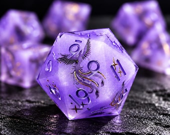 May include: A translucent purple twenty-sided die featuring a gold phoenix graphic and gold numbering. The die has a shimmering, glitter-like internal texture and is surrounded by several other matching purple dice in the background.