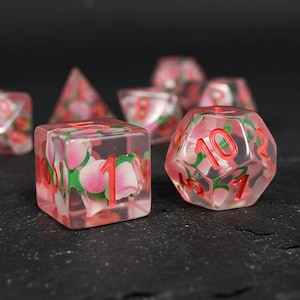 May include: A set of clear resin dice with pink flower inclusions and red numbering. The dice include a variety of shapes, including a cube, a d20, and a pyramid. The dice are designed for tabletop gaming.