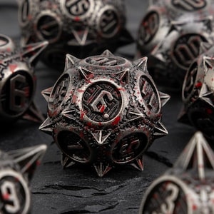 May include: Close-up of a set of metallic dice with a spiked design. The dice are silver with red accents, featuring numbers and intricate patterns. The dice are arranged on a dark, textured surface.