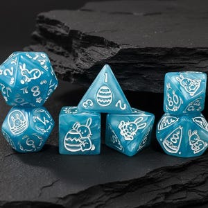 May include: A set of six blue dice with white accents. Each die features a unique design, including numbers, Easter eggs, and bunny illustrations. The dice are arranged on a dark, textured surface.