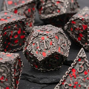 Könnte beinhalten: Ein Set aus polyedrischen Würfeln aus dunkel-silbernem Metall mit detaillierten Drachen-Reliefs und leuchtend roten Farbspritzern. Jeder Würfel hat erhabene Zahlen und einen strukturierten Rand, wobei jede Seite ein aufwendiges Design zeigt.