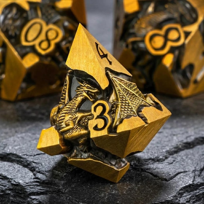 May include: A four-sided die featuring a metallic gold finish with a dark, recessed background. The die displays a sculpted dragon figure with wings spread, along with the engraved numbers 3 and 4 in a bold, black-filled font style.