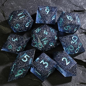 May include: A set of nine polyhedral dice for tabletop games. The dice are a dark blue with a speckled, galaxy-like appearance. The numbers are teal. The dice are arranged on a dark, textured surface.