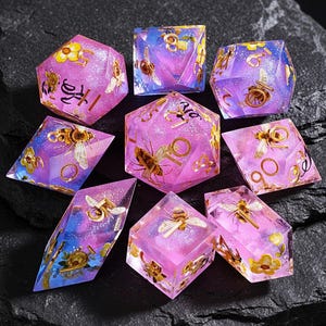 May include: A collection of nine multi-sided dice in pink, purple, and blue hues. Each die features a bee and floral design with gold accents. These dice are for tabletop role-playing games. The dice are measured in centimetres.