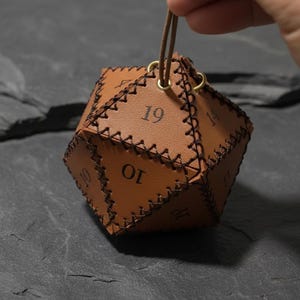 May include: A brown leather die with stitched seams and black numbering. The die is held by a brown cord and has gold-coloured metal eyelets. The numbers 10, 14, 16, and 19 are visible on the faces of the die.