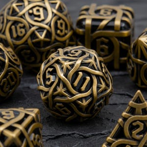 May include: A set of intricately designed dice in various shapes, featuring a metallic gold finish over a black base. The dice have detailed numbers and a complex, interwoven pattern. The image is shot from a close-up angle, highlighting the texture and design.