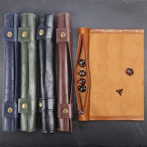 May include: Several leather dice rolling cases in shades of blue, green, black, and brown. Each case is rolled up and fastened with a strap and a brass button. A tan dice bag with a zip and dice is also visible.