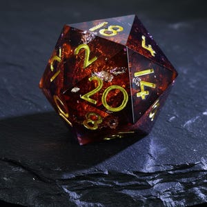 Può includere: Un dado a venti facce con interno marrone rossiccio e numeri dorati. Il dado ha un esterno trasparente e sfaccettato con accenti di foglia d'oro. I numeri sono grandi e facili da leggere, ideale per giochi da tavolo.
