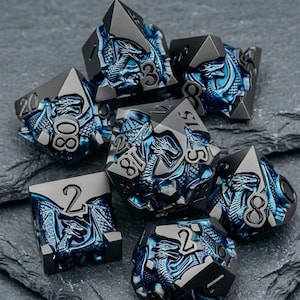 May include: A set of seven polyhedral dice featuring a metallic gunmetal finish with deep blue recessed dragon motifs. Each die displays raised black numbers, including a twenty-sided die, a twelve-sided die, and various other geometric shapes.