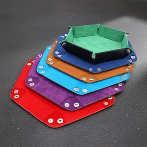 May include: A stack of colorful, hexagonal dice trays in red, purple, blue, brown, and green. Each tray is made of a suede-like material with a contrasting leather trim and metal snap closures. The top tray has a green interior.