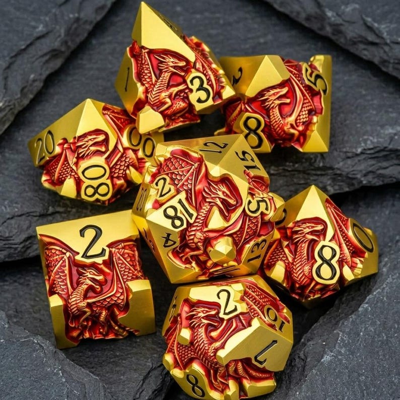 May include: A set of seven polyhedral dice featuring a metallic gold finish with recessed red dragon motifs and black numerals. Each die displays intricate dragon scales and wings, with numbers clearly engraved on the smooth, polished gold surfaces.