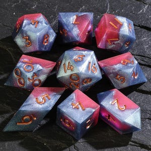 May include: A set of nine polyhedral dice in various shapes. Each die is a mix of blue and red with a glittery effect, and features copper-colored numbers. The dice are designed for tabletop role-playing games.