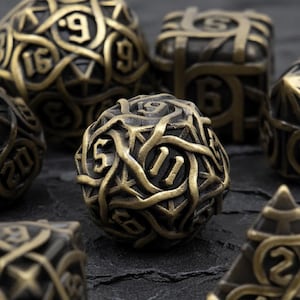 Puede incluir: Primer plano de un juego de dados poliédricos de color bronce. Los dados tienen un diseño intrincado e entrelazado con números claramente visibles. Los dados están dispuestos sobre una superficie oscura y texturizada, adecuada para juegos de mesa.