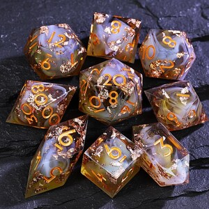 May include: A set of nine polyhedral dice with gold-colored numbers and gold leaf inclusions. The dice are various shapes, including D4, D6, D8, D10, D12, and D20. The dice have a translucent, marbled appearance with shades of brown, white, and yellow.