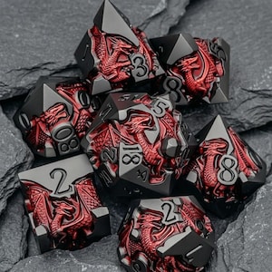 May include: A set of seven polyhedral dice featuring a matte black finish with metallic red dragon reliefs integrated into each face. The numbers are engraved in a clear, bold font, contrasting against the dark metal surfaces of the dice.