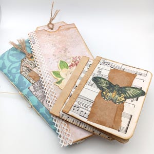 May include: A collection of handmade paper crafts, including a large tag with floral and lace details, a blue patterned journal, and a small notebook with sheet music and a butterfly illustration. The items are tied together with twine.