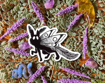 May include: A black and white sticker of a creature with moth-like wings and a feathery tail. The sticker is surrounded by purple lavender, amethyst crystals, a blue snake figurine, and a yellow crescent moon on a bed of green foliage.