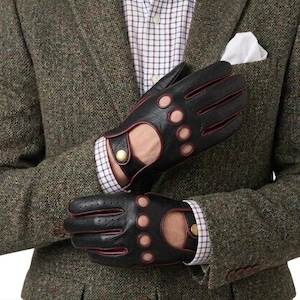 Op de afbeelding: Zwarte leren rijhandschoenen met rode accenten en cirkelvormige uitsparingen. De handschoenen worden gedragen met een tweed jasje en een geruit overhemd. De handschoenen hebben een knoopsluiting bij de pols.