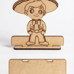 Puede incluir: Un recorte de madera de un niño de dibujos animados con un sombrero y una pajarita. El recorte está de pie sobre una base rectangular con una ranura para un nombre o mensaje. El diseño es de color marrón claro.