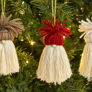 May include: Three handmade Christmas ornaments shaped like gnomes, hanging from gold strings against a green Christmas tree background. Each gnome has a different colored hat and hair, with a cream-colored beard.