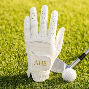 May include: A white golf glove with the initials "AHS" in gold lettering, a golf ball, and a golf club on green grass. The glove has small ventilation holes and a textured palm.