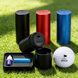 Puede incluir: Juego de regalo temático de golf con una pelota de golf blanca con el logo de una bandera y el nombre "DOUGLAS". Incluye contenedores cilíndricos negros, rojos y azules, una pequeña caja negra con un tubo azul y una botella morada, sobre hierba verde.