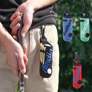 Puede incluir: Soporte negro para pelotas y tees de golf con el nombre "Amelia" en escritura azul. El soporte está sujeto a una trabilla del cinturón con un clip negro. También se muestran soportes adicionales en azul con "Golftatt", verde y rojo.