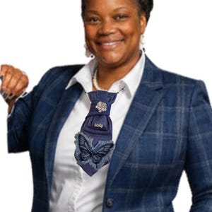 May include: A navy blue denim tie with a butterfly design and a sparkling embellishment. The tie is worn over a white collared shirt and a blue plaid blazer. The butterfly is embroidered on the lower part of the tie.
