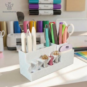May include: A light gray tiered organizer holding various craft tools, including pens, scissors, and weeding tools. The organizer has multiple compartments and is designed to hold Cricut tools. The image also shows rolls of vinyl and Cricut pens.
