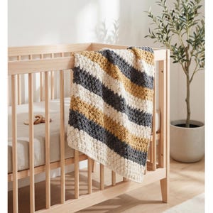 May include: A handmade, striped baby blanket in a crib. The blanket features alternating diagonal stripes of cream, mustard yellow, and gray. The crib is made of light-colored wood and has a neutral-colored mattress.