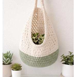Puede incluir: Un macetero colgante de ganchillo en crema y verde salvia. La cesta hecha a mano está llena de pequeñas suculentas verdes. El macetero se suspende de un lazo color crema, perfecto para añadir un toque de naturaleza.