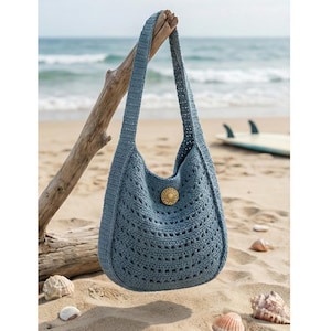 May include: A blue crocheted shoulder bag with a large, decorative button. The bag is hanging from a piece of driftwood on a sandy beach with the ocean in the background. The bag has a textured, open weave design.