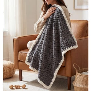 May include: A gray crocheted blanket with a cream-colored border. The blanket is folded and held by a person. The blanket is made of a thick yarn and has a textured appearance. The blanket is displayed on a brown leather chair.