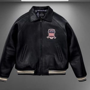 Pode incluir: Um casaco de couro preto com fecho frontal completo, gola clássica e punhos e bainha canelados com riscas brancas. No peito esquerdo, há um emblema que exibe o texto AVIREX acima de um escudo com o design da bandeira americana.