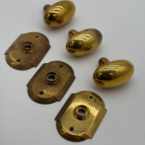 Op de afbeelding: Een verzameling vintage messing deurknoppen en achterplaten. De knoppen zijn ovaal van vorm met een gepolijste gouden afwerking. De achterplaten zijn rechthoekig met afgeronde hoeken en een bijpassende messing afwerking. De hardware ligt op een witte ondergrond.