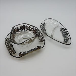 Op de afbeelding: Glazen serveerschalen met zilveren bloemmotieven. De set bevat een vierkante schaal met een kom en een verdeelde ovale schaal. De randen zijn zwart omlijnd. Een vintage serveerset.