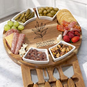 May include: A wooden charcuterie board with a circular serving area and a lower section for cheese knives and a fork. The serving area has white bowls filled with olives, nuts, and other snacks. The board is engraved with "MR. & MRS. WILLIAMS".