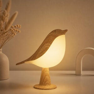 以下が含まれることがあります： 温かい光を放つ、木製の鳥の形をしたテーブルランプ。明るい色の木製のボディと丸みを帯びた、照らされたお腹が特徴です。ランプは木製の台座にあり、自然で魅力的な美しさを生み出しています。