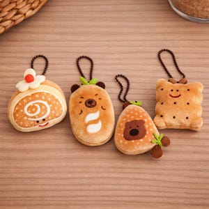 Könnte beinhalten: Vier Plüsch-Schlüsselanhänger in Form einer Biskuitrolle, eines Bären, einer Erdbeere und eines Kekses. Die Schlüsselanhänger sind braun, beige und weiß mit gestickten Details und Kettenschlaufen zum Aufhängen.