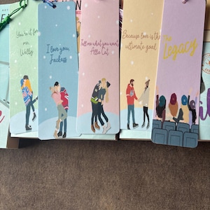 May include: Five rectangular paper bookmarks with tassels, each featuring unique illustrations of couples or groups and printed text including "You're it for me, Wellsy," "I love you, Jackass," "Tell me what you want Allie Cat," "Because love is the ultimate goal," and "The Legacy."