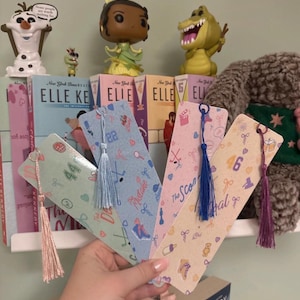 May include: Four rectangular paper bookmarks with rounded corners, each featuring a unique pastel color palette and a distinct tassel. The designs include various small graphic icons like sports equipment, numbers, and text, paired with matching silk-like tassels in light pink, light blue, dark blue, and deep purple.