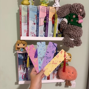 May include: Four rectangular paper bookmarks with colourful patterns and tassels, held in a hand. The bookmarks feature pastel shades of pink, blue, purple, and yellow, decorated with small icons like hockey sticks, skates, and hearts, with text including The Chase, The Risk, The Play, and The Dare.