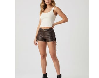 Op de afbeelding: Een vrouw draagt een crèmekleurige tanktop en bruine leren shorts met vetersluiting en metalen studs. Ze draagt ook zwarte enkellaarsjes met een blokhak. De shorts zijn donkerbruin.