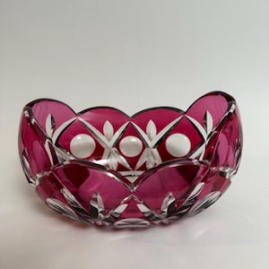 May include: A ruby-coloured, cut-glass bowl with a scalloped rim. The bowl features a geometric pattern of diamond shapes and white circular accents. The bowl is likely used for decorative purposes or serving.