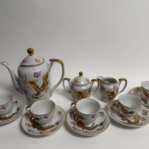 Op de afbeelding: Een wit porseleinen theeservies met gouden accenten en drakenmotieven. De set bevat een theepot, suikerpot, melkkan en zes kopjes met schotels. Het drakenontwerp is goud en rood.