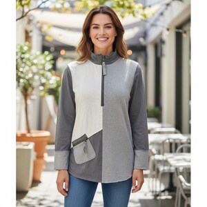 Peut inclure: Haut à manches longues bicolore gris et blanc cassé, avec un col zippé et une petite poche. Le haut a une coupe décontractée et est fait d'une matière douce et confortable. Le modèle porte un jean bleu.