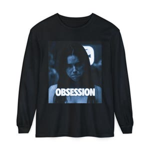 Puede incluir: Una camiseta negra de manga larga con un estampado gráfico cuadrado centrado. El estampado muestra una imagen fotográfica en tonos azules de una persona con cabello largo y oscuro, con la palabra OBSESSION en letras blancas.